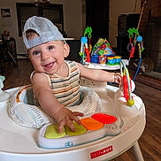 Sammy is registered to the contest to win money with this photo: baby, child, smiling, cap, mesh_cap, striped_shirt, activity_center, toy, piano_keys, indoor, living_room, wooden_floor, furniture, playtime, happy, person, infant, cute, colorful, baby_toys