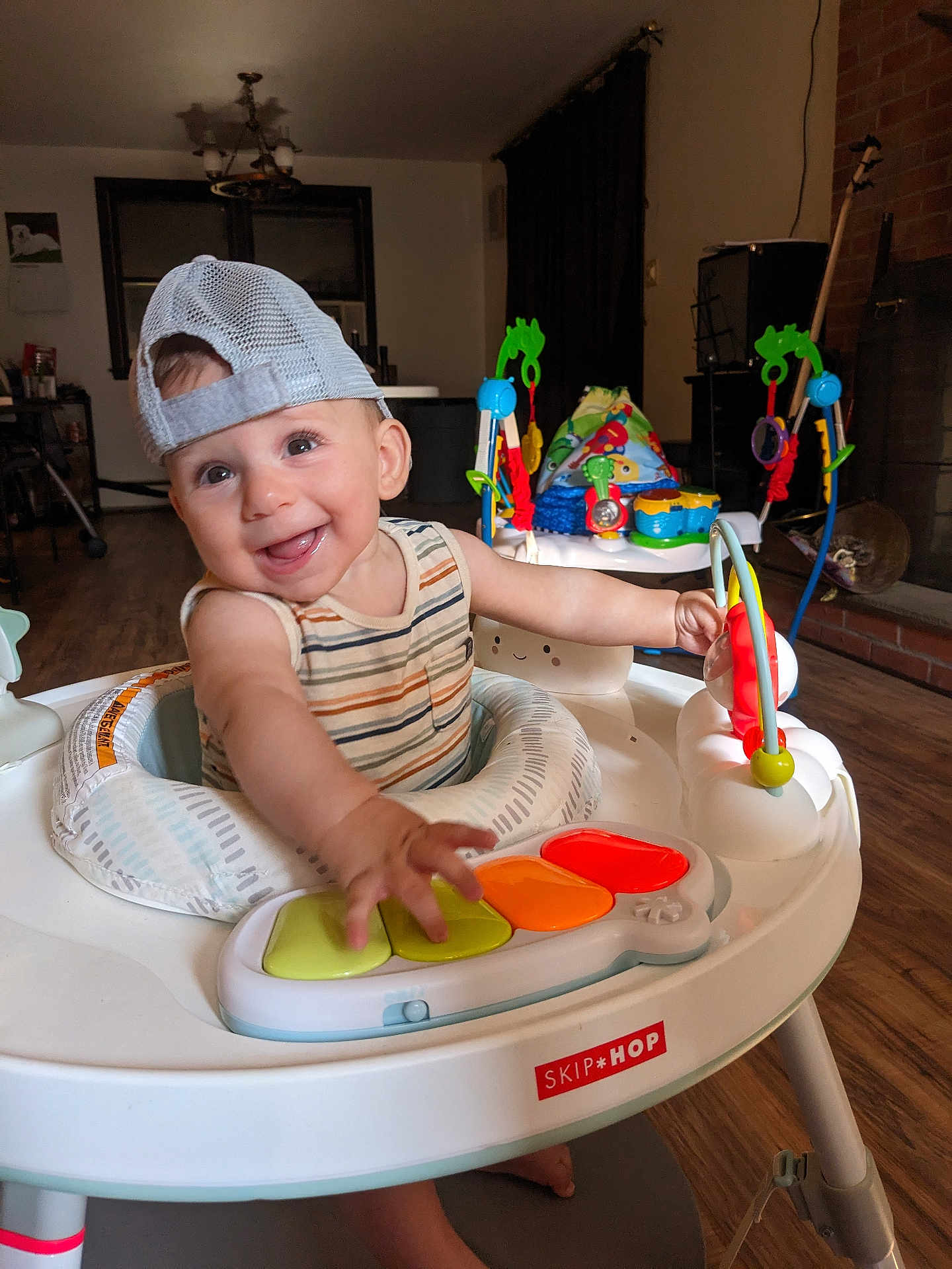 Sammy is registered to the contest to win money with this photo: baby, child, smiling, cap, mesh_cap, striped_shirt, activity_center, toy, piano_keys, indoor, living_room, wooden_floor, furniture, playtime, happy, person, infant, cute, colorful, baby_toys