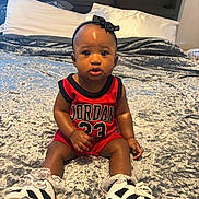 Symphony is registered to the contest to win money with this photo: adorable, baby, basketball_jersey, bed, bedroom, blanket, bow, child, furniture, headband, indoors, infant, jordan_jersey, looking_at_camera, patterned_bedding, pillow, portrait, shoes, sitting, sneakers