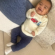 Eiden is registered to the contest to win money with this photo: baby, smiling, child, couch, pillow, white_socks, blue_pants, cream_sweater, cartoon_cars, happy, indoor, cozy, portrait, person, infant, cute, soft_fabric, relaxed, seated, young_child