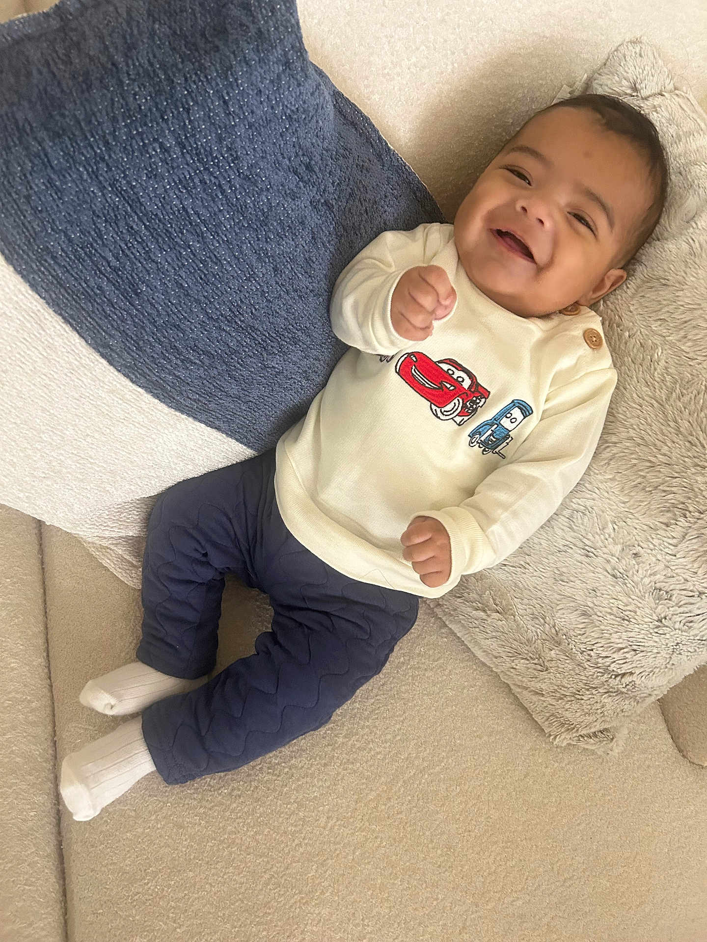 Eiden is registered to the contest to win money with this photo: baby, smiling, child, couch, pillow, white_socks, blue_pants, cream_sweater, cartoon_cars, happy, indoor, cozy, portrait, person, infant, cute, soft_fabric, relaxed, seated, young_child