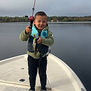 Jaylen is registered to the contest to win money with this photo: child, boat, fishing_rod, fish, water, lake, outdoor, nature, clothing, life_jacket, happy, boy, footwear, sky, trees, recreation, catch, smile, standing, adventure