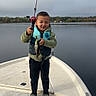 child, boat, fishing_rod, fish, water, lake, outdoor, nature, clothing, life_jacket, happy, boy, footwear, sky, trees, recreation, catch, smile, standing, adventure