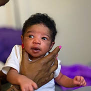 Jeremy joined the competition — help win amazing prizes! adult_hand, baby, blurred_background, bracelet, closeup, cuddling, curly_hair, expression, eyes, hand, holding, indoor, infant, newborn, onesie, pink_nails, portrait, purple_blanket, soft_lighting, tattoo