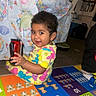 Cecelia is registered to the contest to win money with this photo: toddler, child, smiling, colorful_clothing, play_mat, toy, indoors, floor, carpet, tablecloth, furniture, home, person, happy, young_child, cute, sitting, bright_colors, patterned_clothing, room