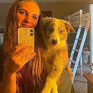 Dexter is registered to the contest to win money with this photo: woman, puppy, dog, selfie, indoor, long_hair, smile, bunk_bed, bedroom, phone, hand, casual_clothing, furniture, carpet, warm_lighting, young_adult, holding, pet, cute, portrait
