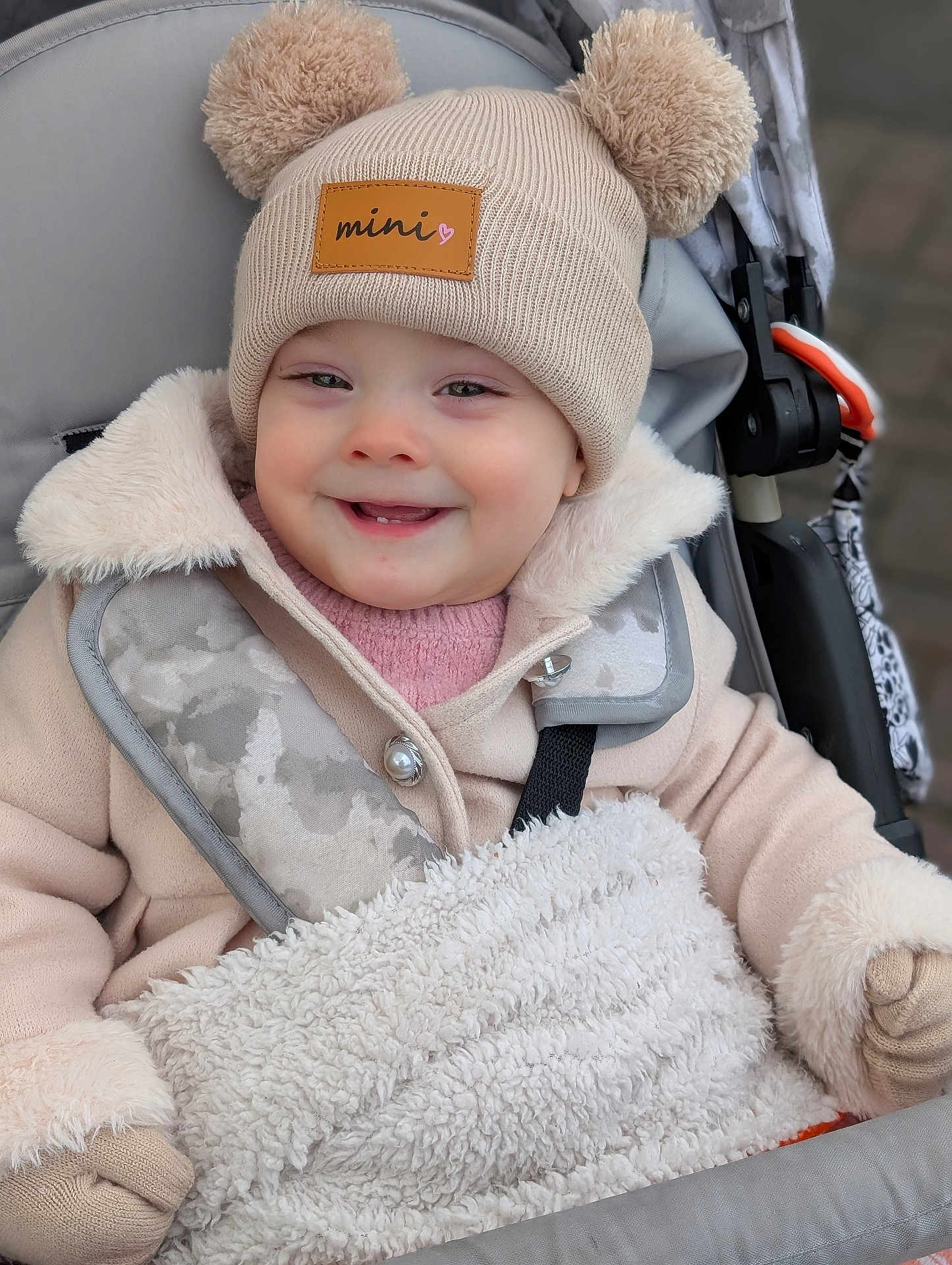 Eloise joined the competition — help win amazing prizes! baby, child, smiling, hat, beige, pom_poms, mittens, coat, stroller, blanket, fuzzy, warm_clothing, outdoor, cute, face, person, winter, comfort, bundle_up, happy