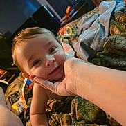 Luca joined the competition — help win amazing prizes! baby, child, hand, smile, face, blanket, camouflage, indoor, toy, curly_hair, person, skin, arm, bed, comfort, cute, expression, infant, portrait, warmth