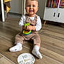 Théo participe au concours pour gagner de l'argent avec cette photo : baby, child, smiling, toy, floor, white_socks, overalls, milestone_card, wooden_floor, bookshelf, books, towel, indoor, person, face, couch, cabinet, happy, sitting, clothing