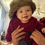 Ellis joined the competition — help win amazing prizes! baby, child, infant, smiling, happy, portrait, fuzzy_hat, winter_clothing, burgundy_outfit, held_by_adult, adult_hands, indoor, living_room, ceiling_fan, window, couch, blanket, close_up, baby_fashion, photography