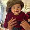 baby, child, infant, smiling, happy, portrait, fuzzy_hat, winter_clothing, burgundy_outfit, held_by_adult, adult_hands, indoor, living_room, ceiling_fan, window, couch, blanket, close_up, baby_fashion, photography