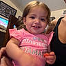 Lailah is registered to the contest to win money with this photo: child, toddler, smiling, portrait, indoors, family, adult, lap, pink_shirt, sparkles, short_sleeves, brown_hair, blue_eyes, snack, sitting, chair, television, living_room, candid, happy