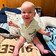 Charles is registered to the contest to win money with this photo: baby, child, smiling, sitting, bed, blanket, pillow, clothing, onesie, indoor, wooden_wall, happy, cute, face, person, infant, foot, hand, crocodile_print, room