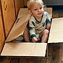 toddler, child, cardboard_box, pajamas, indoor, wooden_floor, kitchen, cabinet, floor, young_child, sitting, playful, blurry, cute, person, home, casual, relaxed, small_child, light