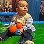 toddler, child, playground, indoor, ball, orange_ball, blue_ball, green_mat, jeans, striped_shirt, knit_sweater, sitting, playing, curious, soft_focus, background, people, colorful, casual_clothing, young_child