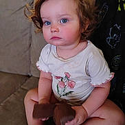 Iris is registered to the contest to win money with this photo: baby, barefoot, blue_eyes, child, couch, curly_hair, cute, flower_print, furniture, home, indoor, person, portrait, serious_expression, sitting, skin, toddler, toy, white_shirt, young_child