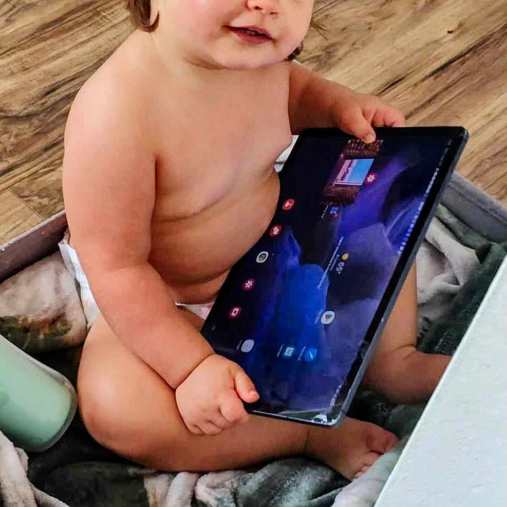 Amaria is registered to the contest to win money with this photo: toddler, child, baby, tablet, technology, indoor, blanket, basket, wood_floor, curly_hair, smile, cute, naked, skin, furniture, play, young_child, person, sitting, curious