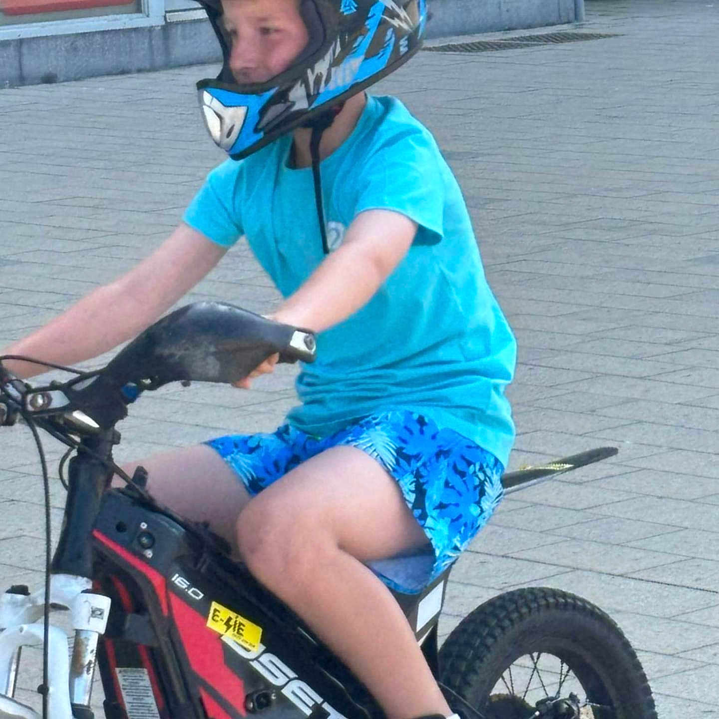 Andgel participe au concours pour gagner de l'argent avec cette photo : bike, black_shoes, blue_shirt, blue_shorts, casual_clothing, child, daylight, electric_bike, glass_windows, helmet, motion, outdoor, pavement, person, protective_gear, reflection, riding, sport, urban, vehicle