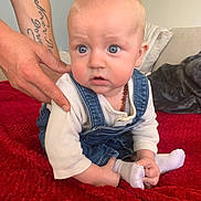 Lénao participe au concours pour gagner de l'argent avec cette photo : baby, child, infant, denim_overalls, white_shirt, tattoo, hand, red_blanket, couch, cushion, indoor, person, sitting, cute, blue_eyes, sock, holding_foot, expression, support, soft_furniture
