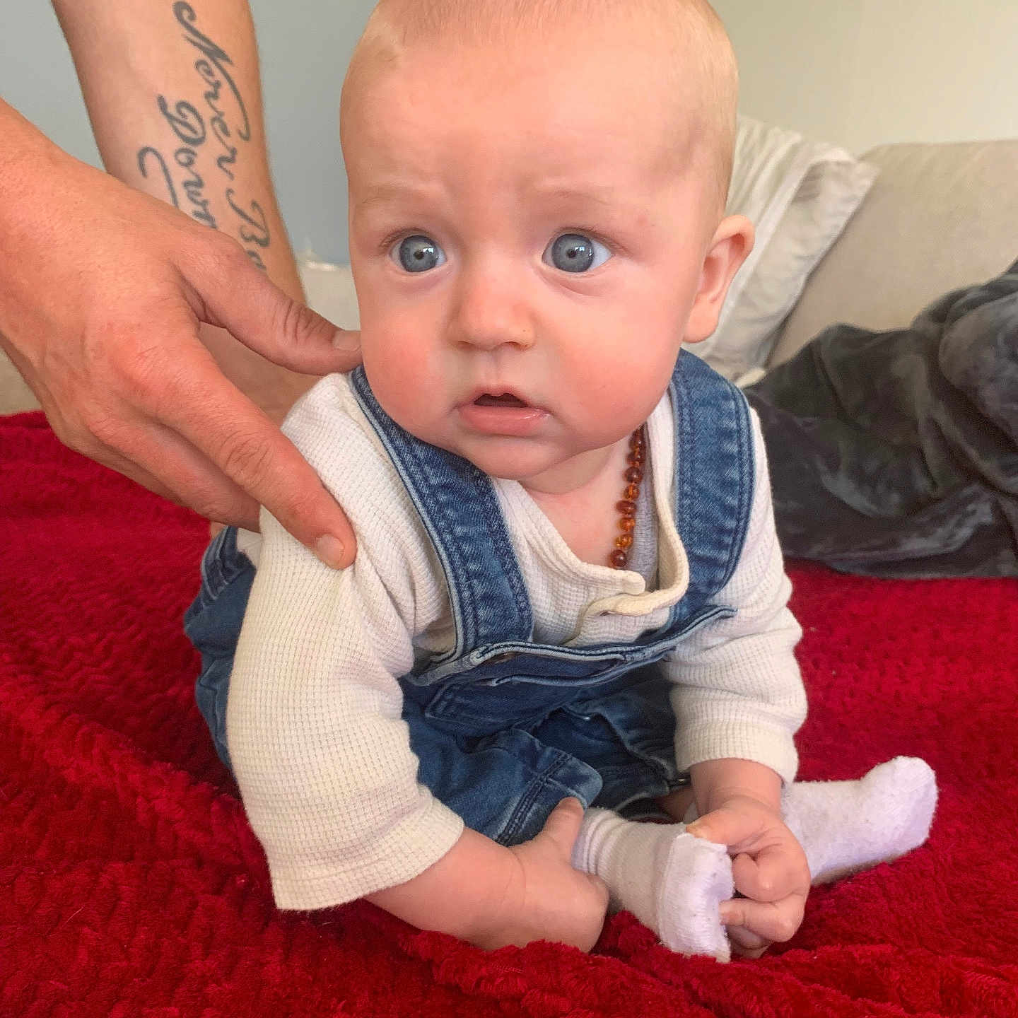 Lénao participe au concours pour gagner de l'argent avec cette photo : baby, blue_eyes, child, couch, cushion, cute, denim_overalls, expression, hand, holding_foot, indoor, infant, person, red_blanket, sitting, sock, soft_furniture, support, tattoo, white_shirt
