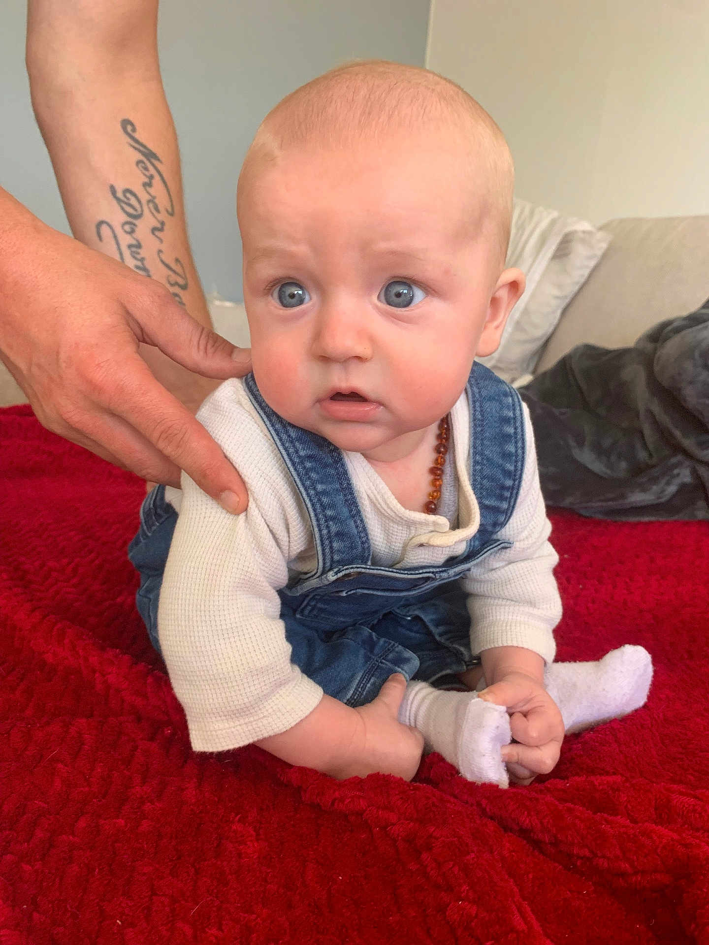 Lénao participe au concours pour gagner de l'argent avec cette photo : baby, child, infant, denim_overalls, white_shirt, tattoo, hand, red_blanket, couch, cushion, indoor, person, sitting, cute, blue_eyes, sock, holding_foot, expression, support, soft_furniture