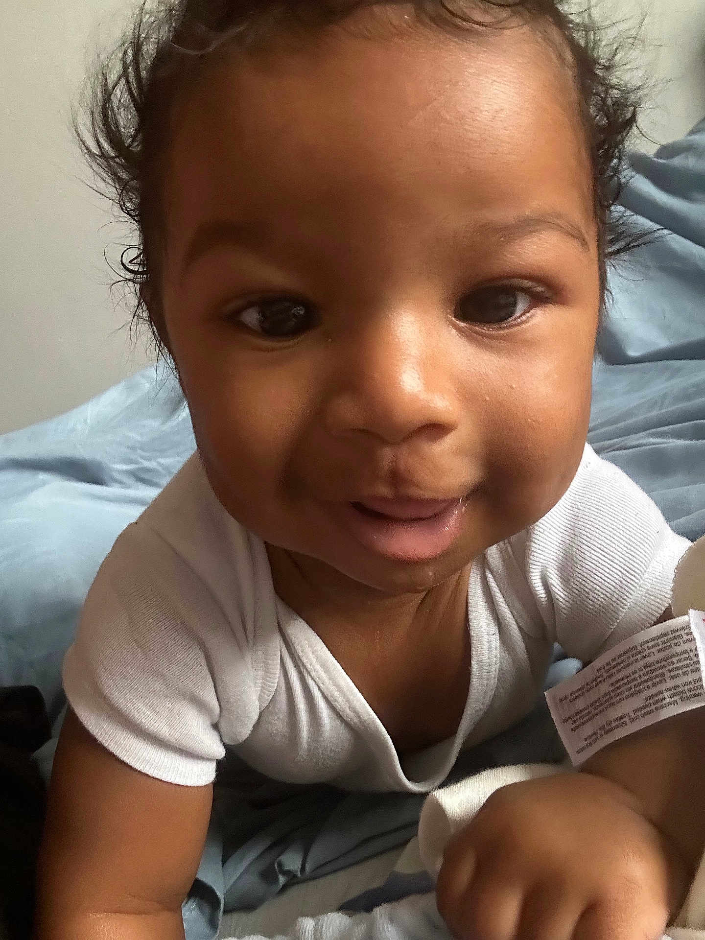Adrien is registered to the contest to win money with this photo: baby, infant, child, smile, face, eyes, white_clothing, onesie, curly_hair, skin, cute, close_up, portrait, happy, lying_down, blanket, soft_texture, indoors, young_child, human