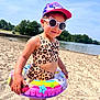 child, beach, sandy_beach, lake, swimsuit, inflatable_ring, sunglasses, hat, summer, outdoor, sunny, water, trees, cute, smiling, fashion, playful, vacation, fun, person