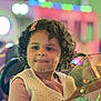 child, curly_hair, pink_hair_clip, vest, long_sleeve_shirt, amusement_ride, glass_windshield, colorful_lights, bokeh, face, tongue_out, playful, indoor, portrait, seat, smiling, cute, young, person, fun