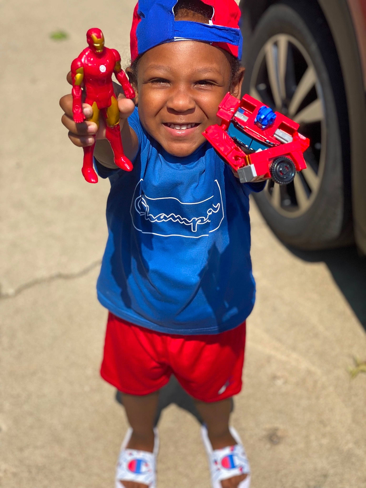 Maddux is registered to the contest to win money with this photo: arm, automotive_tire, baseball_cap, cap, car, electric_blue, eye, face, fun, happy, headwear, organ, people, person, red, shorts, smile, t_shirt, tire, toddler