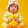 toddler, child, yellow, plush_toy, chicken_hat, cute, soft_texture, indoor, portrait, baby, costume, sitting, orange, yellow_background, toy, funny_hat, young_child, clothing, face, hands