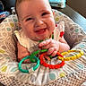 Avilyn is registered to the contest to win money with this photo: baby, smiling, toy, colorful, rings, baby_seat, indoor, happy, child, infant, face, hand, clothing, window, natural_light, pattern, furniture, play, cute, cheerful