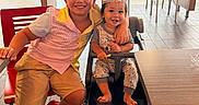 Tom is registered to the contest to win money with this photo: child, toddler, smiling, crown, restaurant, high_chair, red_crocs, shorts, striped_shirt, table, chair, floor, window, daylight, happy, family, indoor, person, cute, playful