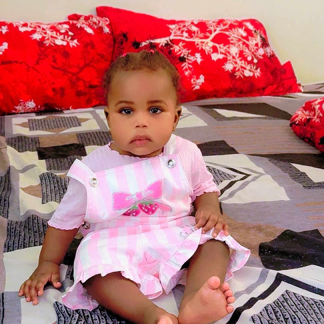 Afrah joined the competition — help win amazing prizes! baby, child, infant, bed, bedding, pillow, red_pillow, patterned_bedspread, pink_dress, striped_dress, bow, barefoot, indoors, sitting, portrait, cute, calm, person, floor_covering, home