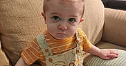 Damon is registered to the contest to win money with this photo: baby, child, couch, cushion, hair, mohawk, orange_shirt, overalls, pooh, seat, indoor, serious_face, person, furniture, living_room, expression, clothing, cute, young_child, home
