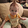baby, child, couch, cushion, hair, mohawk, orange_shirt, overalls, pooh, seat, indoor, serious_face, person, furniture, living_room, expression, clothing, cute, young_child, home