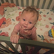 Willow is registered to the contest to win money with this photo: baby, child, infant, crib, crib_rail, floral_bedding, bedroom, indoors, portrait, face, smiling, curious, sitting, standing, hand, striped_shirt, toy, clothing, wooden_furniture, home