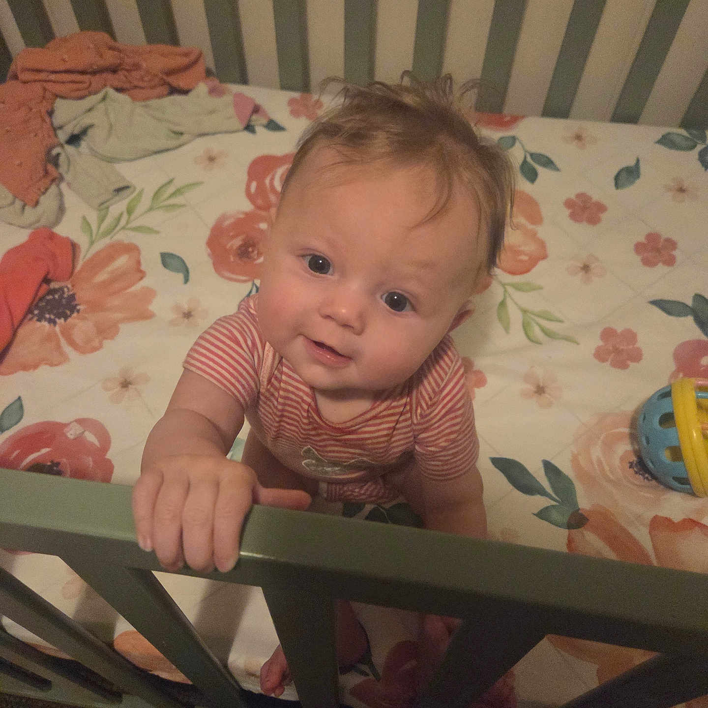 Willow is registered to the contest to win money with this photo: baby, child, infant, crib, crib_rail, floral_bedding, bedroom, indoors, portrait, face, smiling, curious, sitting, standing, hand, striped_shirt, toy, clothing, wooden_furniture, home