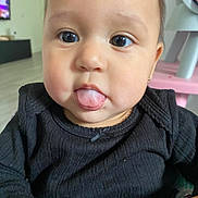 Adalyn is registered to the contest to win money with this photo: baby, child, face, tongue, expression, black_clothing, indoor, close_up, cute, young_child, person, skin, eyes, cheeks, earring, furniture, television, floor, portrait, home