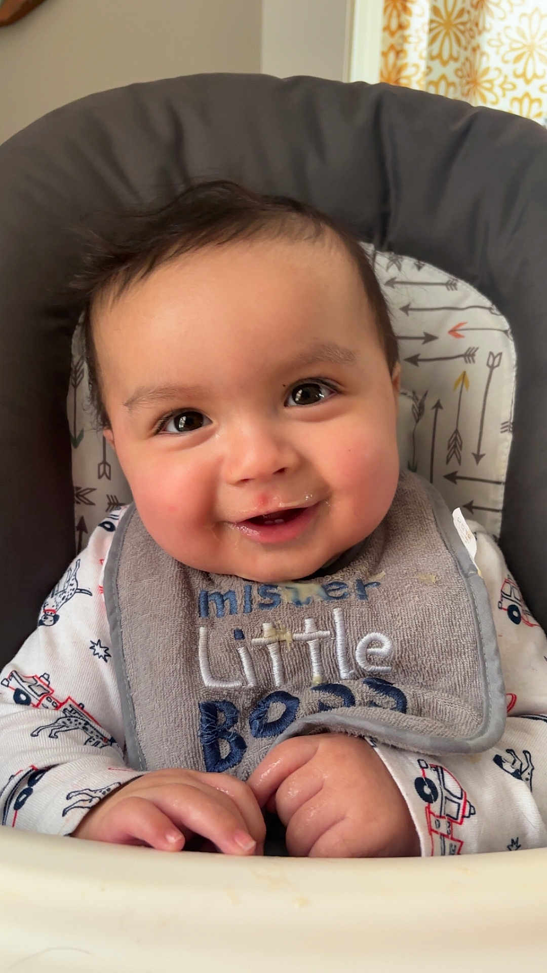 Amir is registered to the contest to win money with this photo: baby, infant, smiling, high_chair, bib, clothing, face, hands, indoor, cute, child, happy, portrait, person, young_child, feeding_chair, patterned_clothing, home, closeup, expression