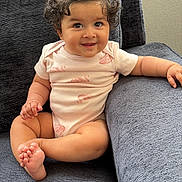 Sakina is registered to the contest to win money with this photo: baby, child, toddler, sitting, smiling, portrait, curly_hair, topknot, pink_outfit, onesie, barefoot, couch, indoor, home, furniture, happy, infant, arms, legs, photography