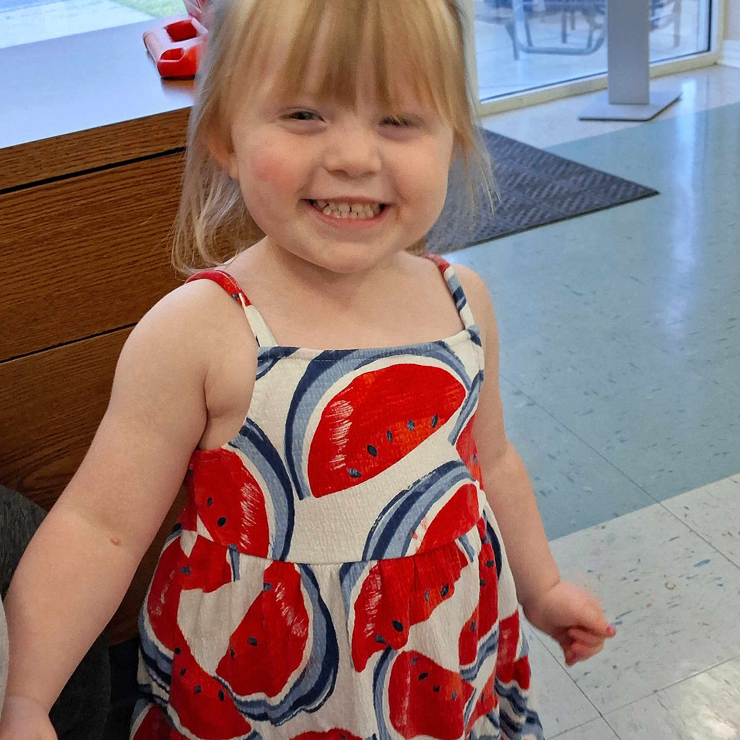 Rylee joined the competition — help win amazing prizes! blonde_hair, casual_clothing, cheerful, child, cute, daylight, dress, floor, girl, happy, indoor, person, playful, portrait, smiling, standing, watermelon_pattern, window, wooden_counter, young_child
