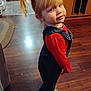 blonde_hair, cabinet, casual_clothing, child, denim_overalls, face, floor, hair, home_interior, indoor, person, pigtails, playful, pose, red_shirt, smile, standing, white_sneakers, wooden_floor, young