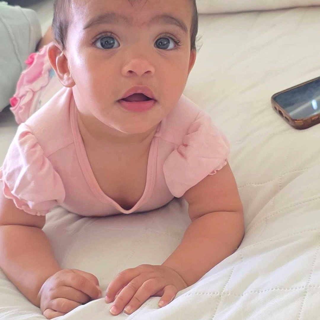 Hazel is registered to the contest to win money with this photo: baby, child, curly_hair, blue_eyes, pink_clothing, bed, quilted_bedspread, smartphone, indoor, portrait, infant, person, cute, lying_down, expression, hands, face, looking_up, soft_lighting, home