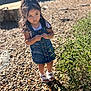 Ashanti is registered to the contest to win money with this photo: child, girl, denim_dress, white_sandals, outdoor, pebbles, green_bush, sunlight, shadow, standing, pensive, young, nature, casual_clothing, summer, daylight, portrait, cute, fashion, plant