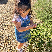 Ashanti is registered to the contest to win money with this photo: child, girl, denim_dress, white_sandals, plant, greenery, sunlight, outdoor, pebbles, nature, curious, exploring, summer, casual_clothing, portrait, daylight, young_child, hair_tied, standing, shoes