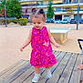toddler, child, dress, pink, outdoor, park, wooden_platform, walking, hair_buns, shoes, sneakers, buildings, trees, cars, sidewalk, daylight, person, playground, urban, happy