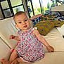 baby, child, infant, sitting, couch, white_leather, floral_dress, blue_eyes, indoor, pillows, patterned_pillows, colorful, window, daylight, home_interior, furniture, cute, portrait, smiling, person