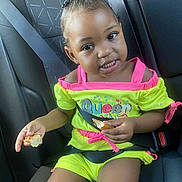 Amira is registered to the contest to win money with this photo: toddler, child, car_seat, seatbelt, eating, food, yellow_clothing, pink_clothing, hair_beads, smiling, indoors, car_interior, happy, person, young_child, cute, portrait, seat_belted, sitting, bright_colors