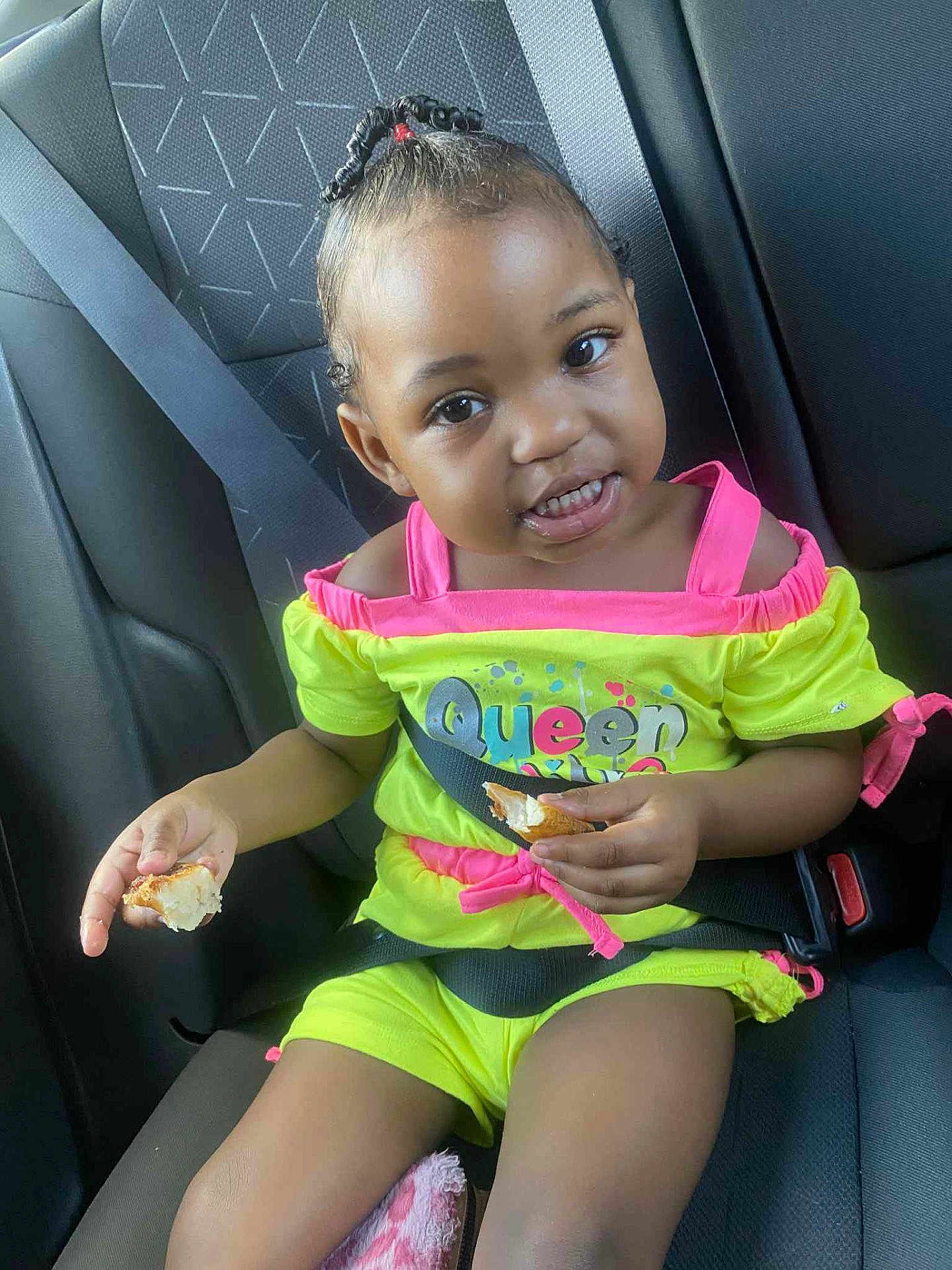Amira is registered to the contest to win money with this photo: toddler, child, car_seat, seatbelt, eating, food, yellow_clothing, pink_clothing, hair_beads, smiling, indoors, car_interior, happy, person, young_child, cute, portrait, seat_belted, sitting, bright_colors