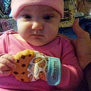 Nova joined the competition — help win amazing prizes! baby, child, pink_clothing, hat, teething_mitten, cookie_pattern, indoor, furniture, curtain, face, hand, person, clothing, expression, seated, room, background, household_items, portrait, serious